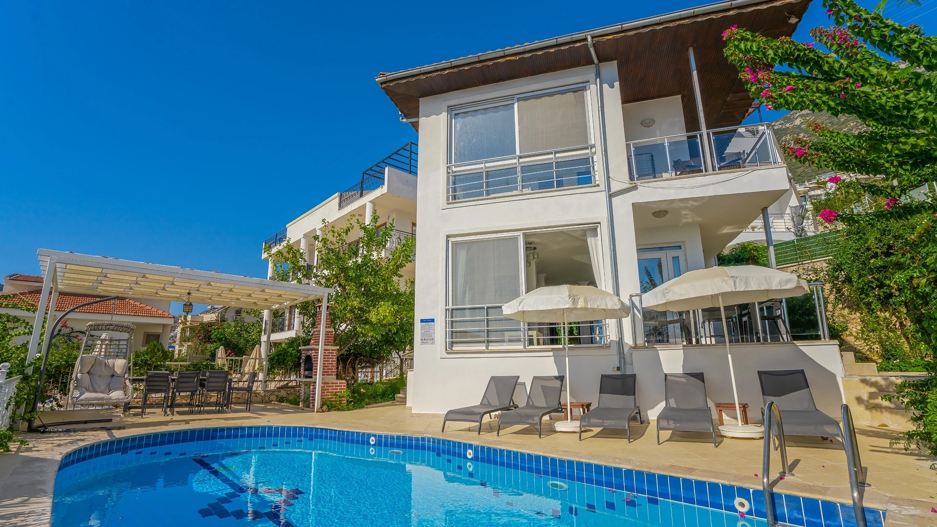 Daytime exterior view of the villa with pool, lounging area, and scenic hillside setting.