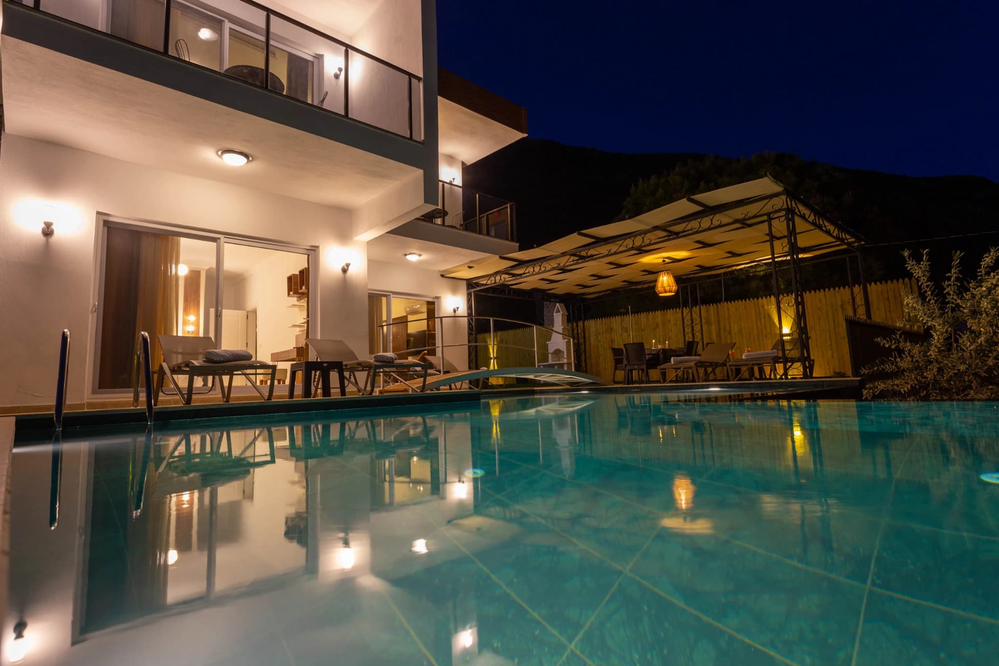 Illuminated private pool with terrace, lounge area, and pergola at night.