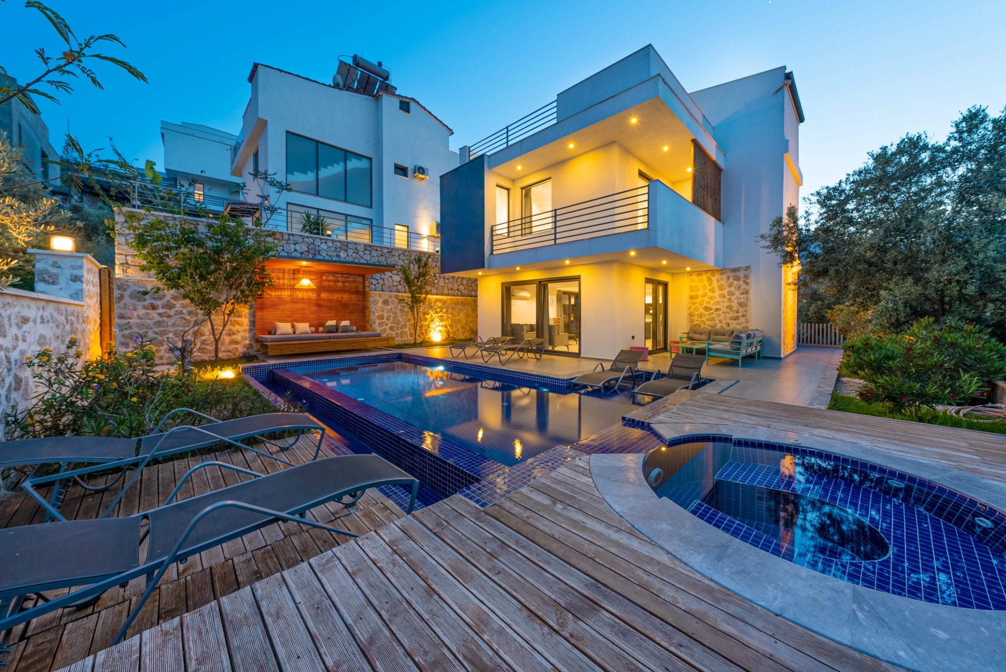 Resort-style pool and jacuzzi area with loungers and illuminated stone walls at twilight.