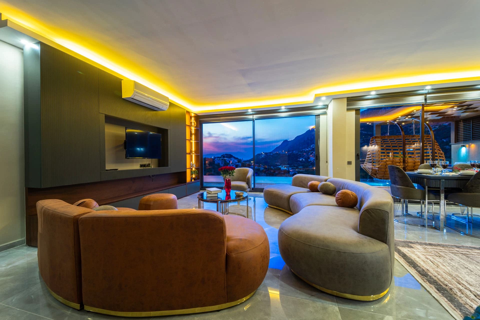 Contemporary living room with curved designer seating, wall-mounted TV, and floor-to-ceiling windows showcasing mountain and valley views at dusk.