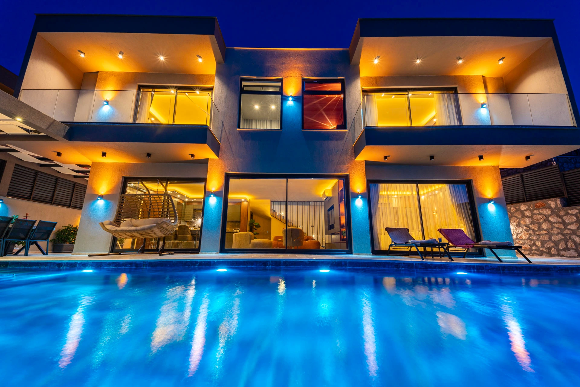 Blue-lit private pool fronting the modern villa with colorful lounge seating.