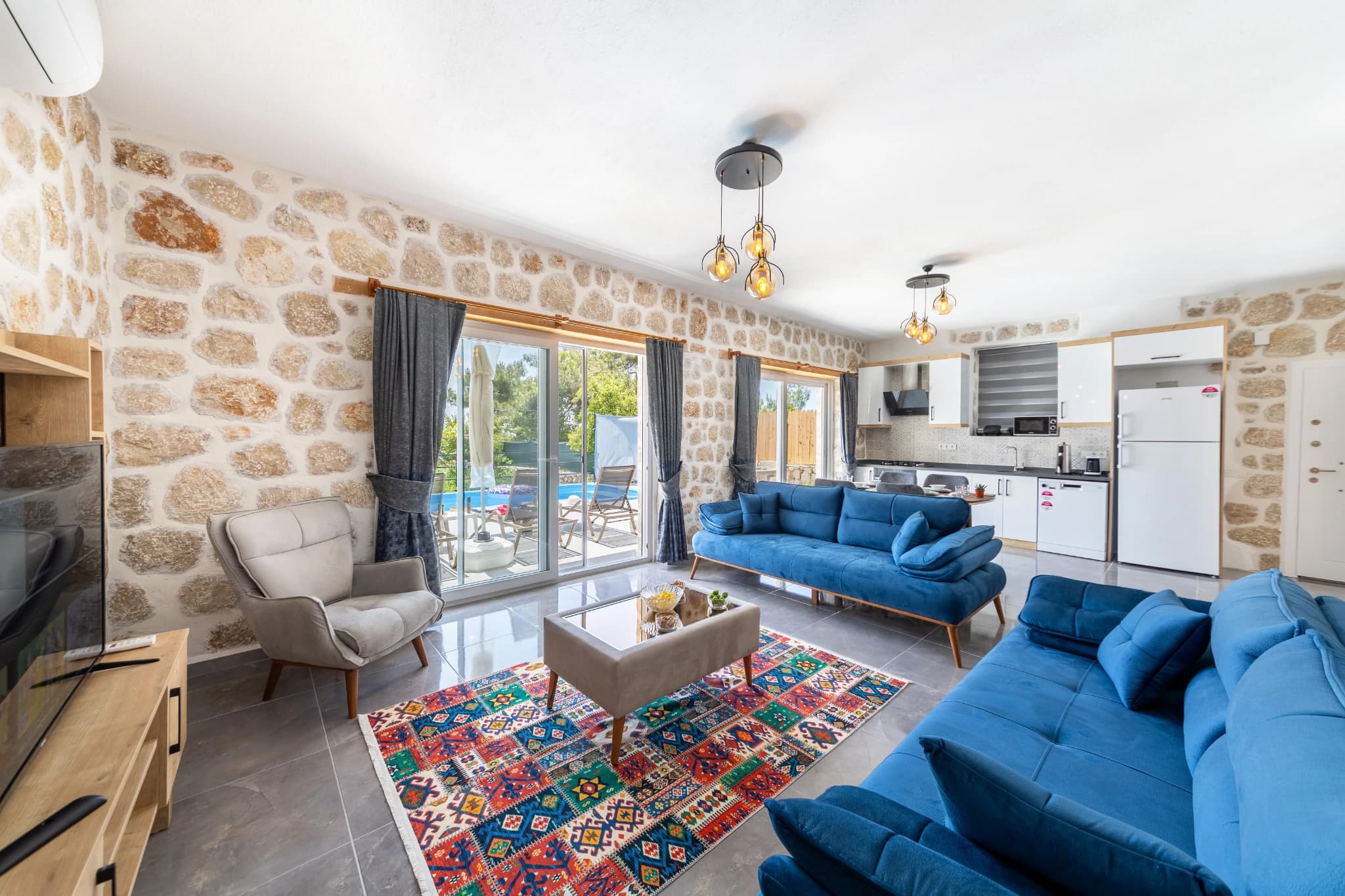 Spacious open-plan living room with blue sofas, stone walls, and pool view through sliding doors.