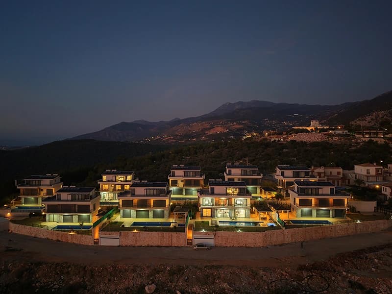 Evening aerial view of the villa complex nestled in mountains with illuminated pools and contemporary design.