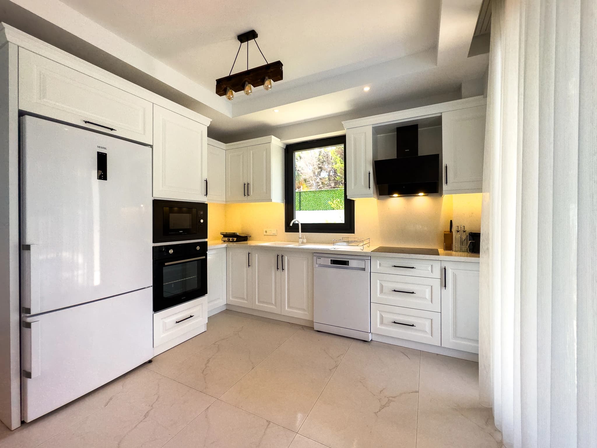 Modern white kitchen with black appliances, yellow accent backlighting, and garden views.