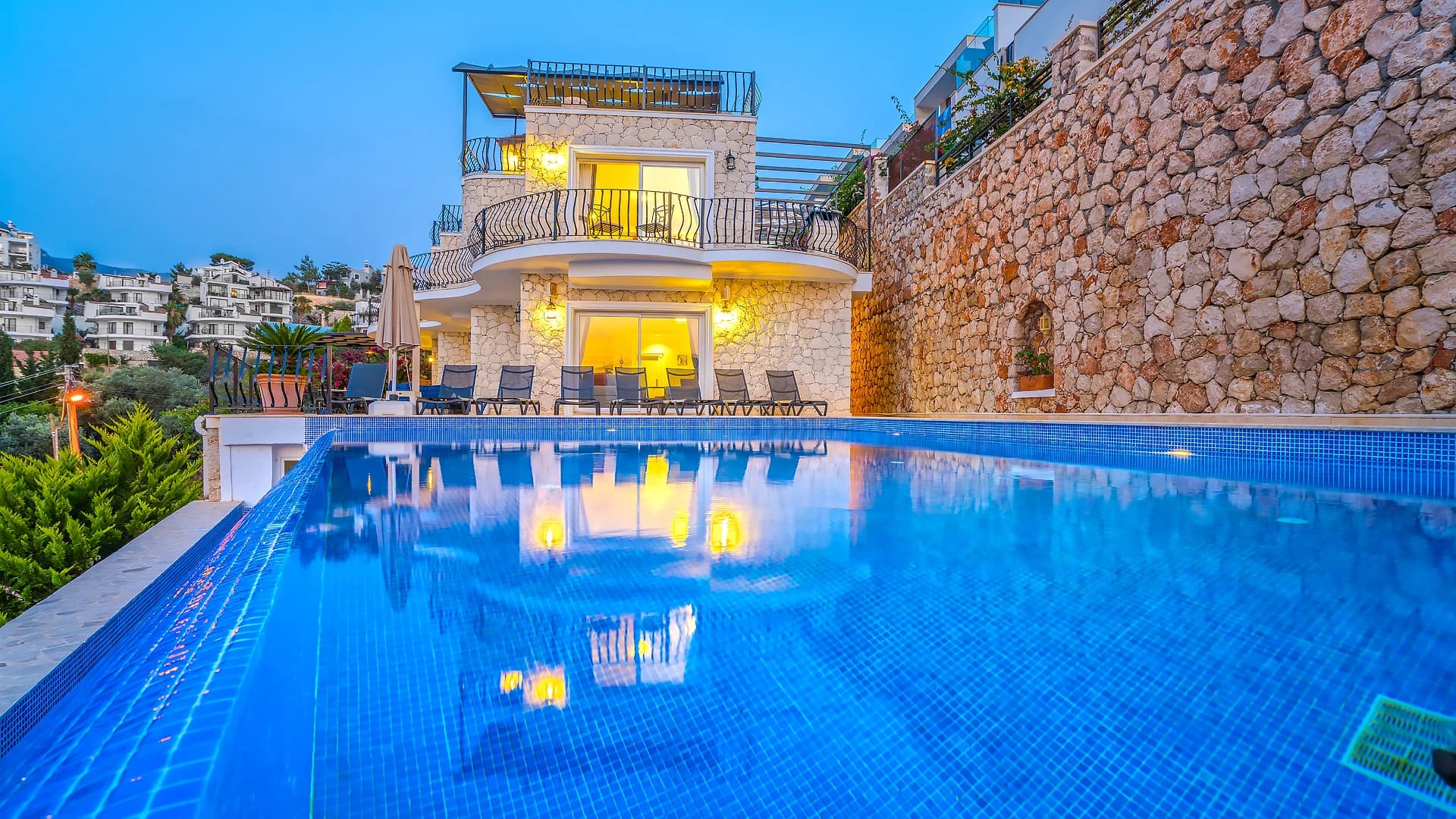 Illuminated private pool with sun loungers overlooking the hillside town at dusk.