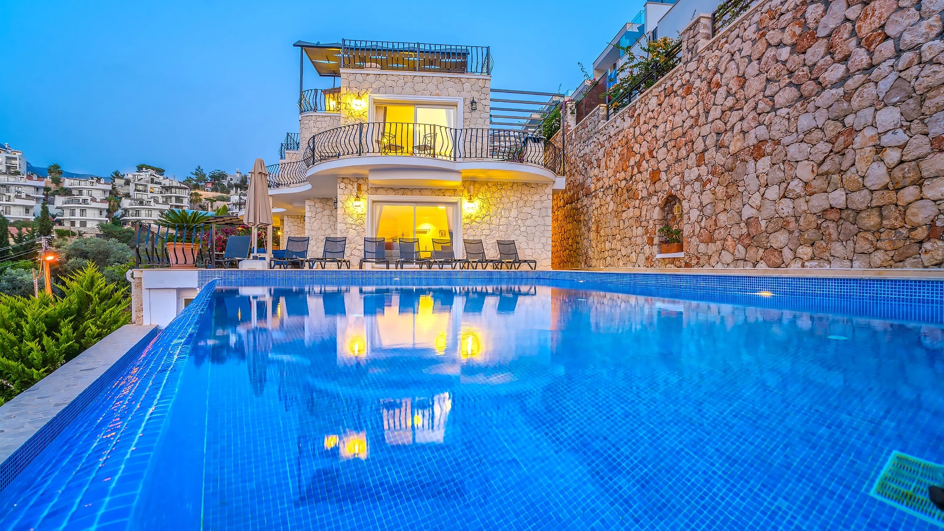 Illuminated private pool with sun loungers overlooking the hillside town at dusk.