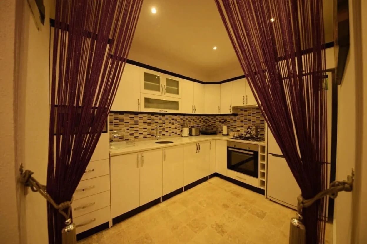 Kitchen view with warm lighting, white cabinetry, and elegant burgundy string curtains.