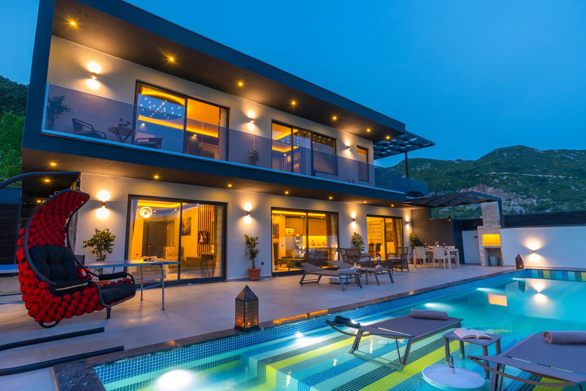 Modern villa exterior with illuminated infinity pool and mountain backdrop at dusk.