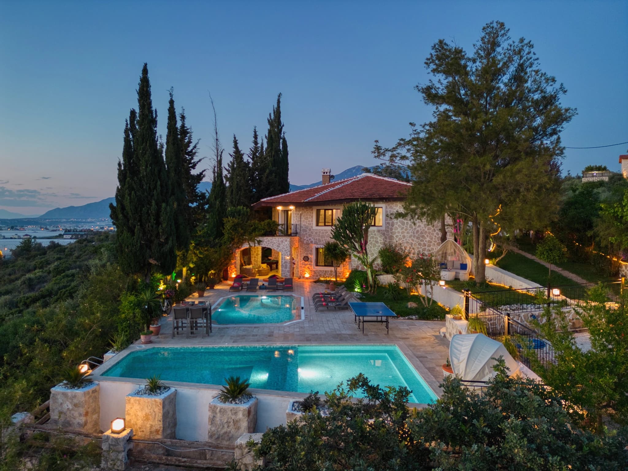 Stunning twilight view of dual pools with sea view, lounging areas, and Mediterranean landscape.