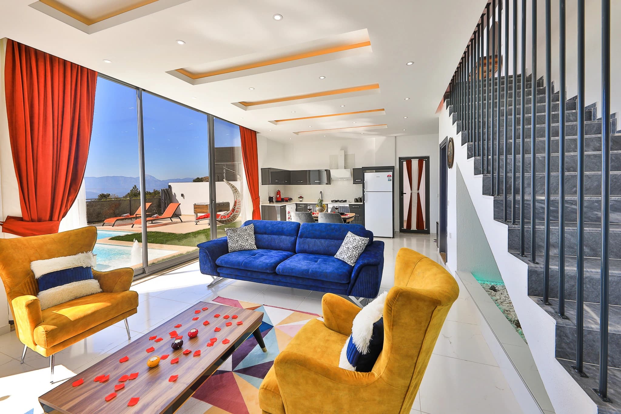 Bright living room with blue sofa, yellow chairs, and panoramic sea and mountain views through floor-to-ceiling windows.