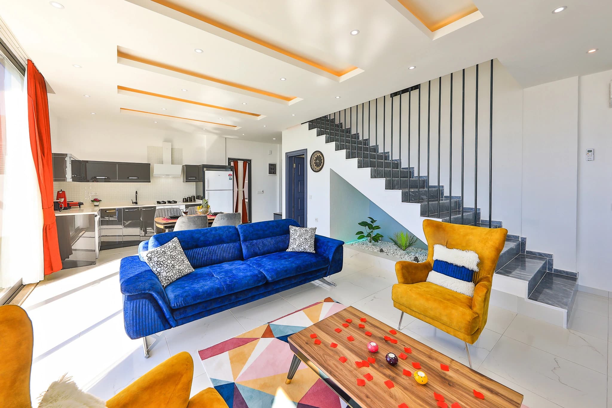Open-plan living area with blue sofa, yellow seating, foosball table, and integrated kitchen with staircase.