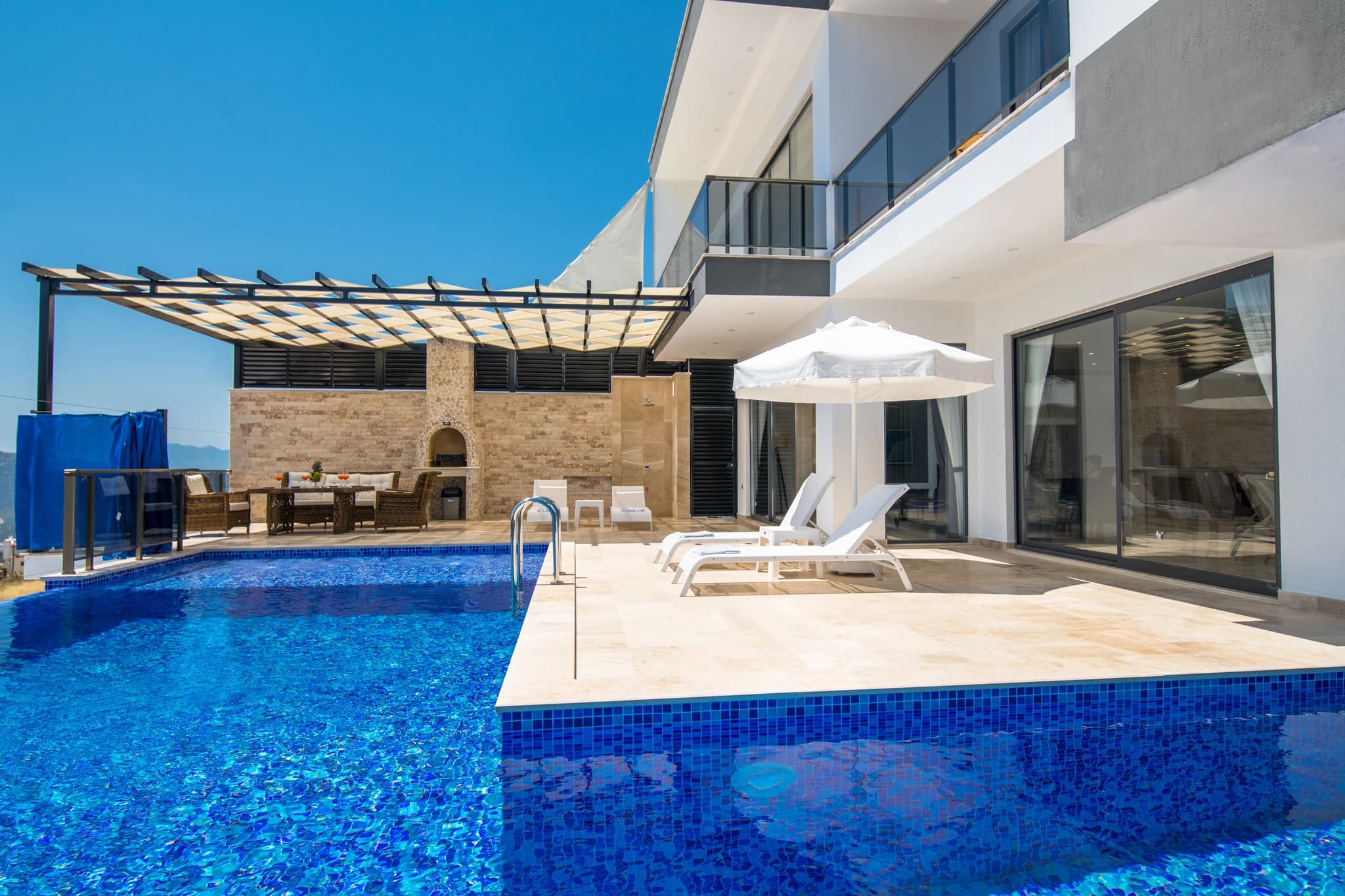 Abgeschiedene Flitterwochen-Villa mit privatem und beheizbarem Innenpool in Kalkan – Resort-style pool area with dual pools, lounge chairs, pergola shade structure, and stone fireplace on expansive terrace.
